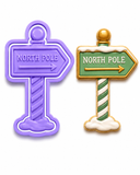 Royal Orchid Lavender North Pole sign cookie cutter and embosser displayed on a white background beside a decorated North Pole sign sugar cookie with green sign, candy cane post, and snowy base."