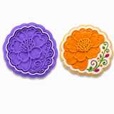 Royal Orchid Lavender marigold flower plaque cookie cutter and embosser displayed beside a decorated sugar cookie featuring a large orange marigold flower with colorful Mexican folk-art floral accents on a white background.