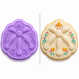 Pan de Muerto cookie cutter and embosser shown beside a decorated sugar cookie inspired by the traditional Mexican Day of the Dead sweet bread with raised bone details and colorful folk art accents.