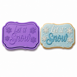 Purple Let It Snow snowflake plaque cookie cutter and embosser stamp set shown beside a decorated blue winter snowflake sugar cookie.